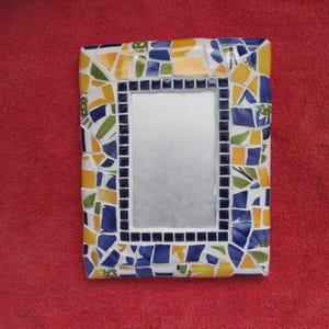 May include: Rectangular mosaic mirror with a colorful border. The frame features a mosaic design with blue, yellow, green, and white tiles. The mirror's inner frame is lined with small black squares, creating a decorative accent.
