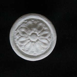 May include: A white, circular decorative element with a floral design. The raised pattern features a central flower surrounded by petal-like shapes. The object is set against a black background, highlighting its texture and form.