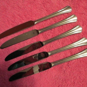 May include: Five silver dinner knives with ornate handles. The knives are laid out on a pink fuzzy surface.