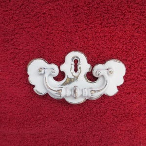 May include: A white metal drawer pull with a distressed finish revealing gold-toned metal underneath. The hardware features a central keyhole escutcheon plate and a curved, swinging handle with ornate, symmetrical scrollwork detailing on both sides.