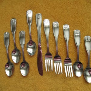 May include: A collection of antique silverware, including spoons, forks, and a knife, arranged on a golden-yellow surface. The silverware has a shiny, reflective surface and a decorative design on the handles. The knife has a dark red blade.