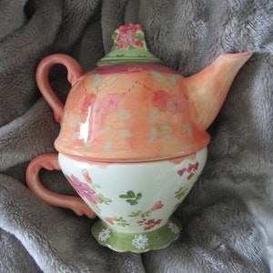 May include: A pink and white floral teapot with a matching teacup. The teapot has a lid with a floral design and a handle. The teacup has a handle and a floral design.