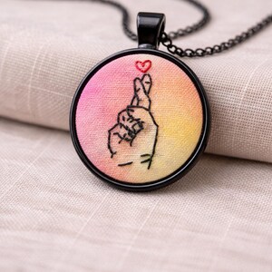 May include: A round pendant necklace with a black chain. The pendant features an embroidered hand gesture with a small red heart at the fingertips, set against a pink and yellow gradient background. The pendant is framed by a black border.