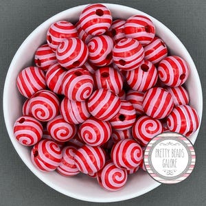 May include: A white bowl filled with red and white striped beads. The beads have a spiral design and are round with a hole through the center. The beads are labeled "Pretty Beads Galore" in a circular logo.