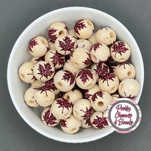 May include: A white bowl filled with round, cream-colored beads. Each bead features a red maple leaf design. The beads have a hole drilled through the center. The bowl is on a gray surface. The image includes a logo that says "Pretty Charms & Beads."