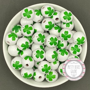 May include: A white bowl filled with white wooden beads, each adorned with a green shamrock design. The beads are round and have a glossy finish. The image includes a circular logo that reads "Pretty Beads Galore".