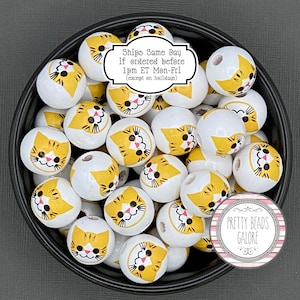 May include: A bowl filled with white wooden beads with yellow and black cat faces painted on them. The beads are for crafting and jewelry making.