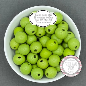 May include: A bowl filled with 20 bright green wooden beads. Each bead has two holes drilled through it. The beads are in a white bowl. A sticker on the bowl says "Ships Same Day if ordered before 1pm ET Mon-Fri (except on holidays)". A sticker on the right side of the bowl says "PRETTY BEADS GALORE".