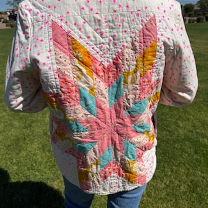 Może przedstawiać: Pikowana kurtka z wzorem rozbłysku gwiazdy na plecach. Kurtka jest biała z różowymi wzorami gwiazd i dużym rozbłyskiem gwiazdy w odcieniach różu, żółci i turkusu. Kurtka jest noszona na niebieskich dżinsach.