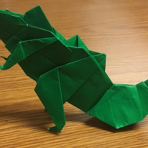 May include: A green origami dinosaur with a long tail and a head that is slightly tilted to the side. The dinosaur is standing on a wooden surface.