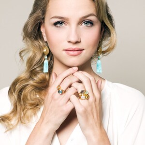 Puede incluir: Una mujer con una camisa blanca con anillos de oro en los dedos. Los anillos presentan varias piedras preciosas, incluyendo azul, verde y marr&oacute;n. Tambi&eacute;n lleva pendientes de oro con borlas azules.