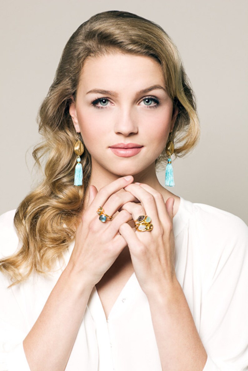 Puede incluir: Una mujer con una camisa blanca con anillos de oro en los dedos. Los anillos presentan varias piedras preciosas, incluyendo azul, verde y marr&oacute;n. Tambi&eacute;n lleva pendientes de oro con borlas azules.