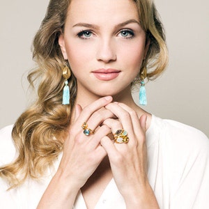 May include: A woman with long blonde hair wearing gold earrings with blue tassels and gold rings with colourful gemstones.