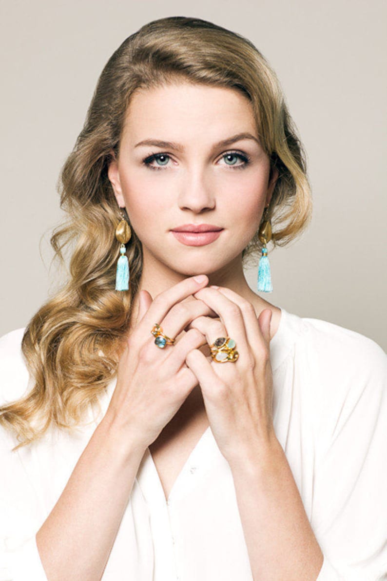 May include: A woman with long blonde hair wearing gold earrings with blue tassels and gold rings with colourful gemstones.