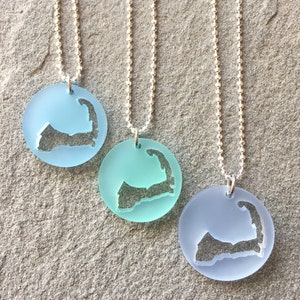 May include: Three silver chain necklaces with frosted glass pendants shaped like the outline of Martha's Vineyard. The pendants are in shades of blue and light blue.