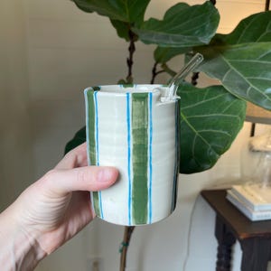 May include: A white ceramic mug with vertical green and blue stripes. The mug has a clear glass straw inserted into it. The mug is cylindrical and appears to be handmade. The background includes a large-leafed plant.