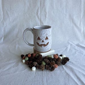 May include: A white ceramic mug with a speckled finish and a brown jack-o'-lantern face design. The mug features a curved handle and sits atop a small white block surrounded by a collection of small brown pinecones and tiny decorative pumpkins.