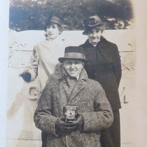 May include: A black and white vintage photograph of three people. Two women are wearing hats and coats. The woman on the left is looking to the side. The woman on the right is looking at the camera. The man in the middle is wearing a hat and coat and holding a camera. The photo is taken outdoors in front of a building.
