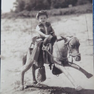 May include: A black and white photo of a young child wearing a cowboy hat and riding a toy horse.