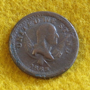 May include: A copper-colored 1863 United States penny coin. The coin features a profile of a person and the words "UNITED STATES OF AMERICA" around the edge. The year "1863" is visible below the profile.