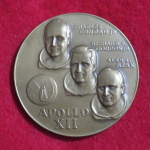 May include: A bronze Apollo XIII commemorative coin. The coin features portraits of Charles Conrad Jr., Richard F. Gordon Jr., and Alan L. Bean. The text "APOLLO XIII" and the mission's logo are also visible.