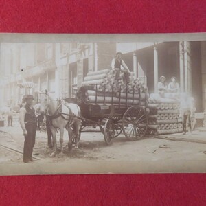 May include: A black and white photo of a horse-drawn wagon loaded with rolled up fabric. The wagon is on a city street with buildings in the background. There are several people standing around the wagon.