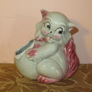May include: A ceramic planter shaped like a gray cat with pink accents. The cat has large eyes and a wide smile. The planter has a hole in the back for drainage.