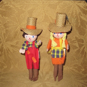 May include: Two handmade dolls, a boy and a girl, are dressed in rustic attire. The boy doll wears red overalls with orange patches and a plaid shirt. The girl doll is in an orange dress with a plaid apron. Both dolls have straw hats.