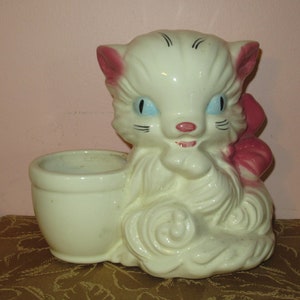 May include: A vintage ceramic planter in the shape of a white cat with pink accents. The cat has blue eyes and a pink bow. The planter has a small, round opening for plants.