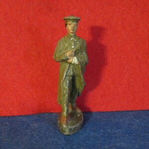 May include: A green painted lead toy soldier figurine wearing a hat and holding a rifle. The soldier is standing on a base.