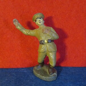 May include: A vintage toy soldier figurine, painted in a light brown color, wearing a cap and a uniform. The soldier is standing with one arm raised, as if giving a salute.