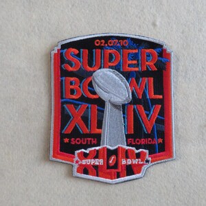 May include: A rectangular embroidered patch featuring the text SUPER BOWL XLIV, 02.07.10, and SOUTH FLORIDA. The design includes a silver Vince Lombardi trophy graphic, red lettering, and blue palm tree silhouettes on a black background with a red border.