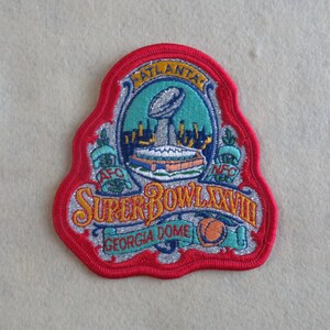 May include: A Super Bowl XXVIII embroidered patch featuring a red border, silver glitter background, and a central graphic of the Vince Lombardi Trophy. The text reads Atlanta, AFC, NFC, Super Bowl XXVIII, and Georgia Dome, with a peach icon.