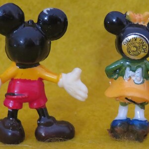 May include: Two small, vintage Mickey Mouse and Minnie Mouse figurines. Mickey is in red shorts, a yellow shirt, and white gloves. Minnie wears a green top and orange skirt. Both have black ears.