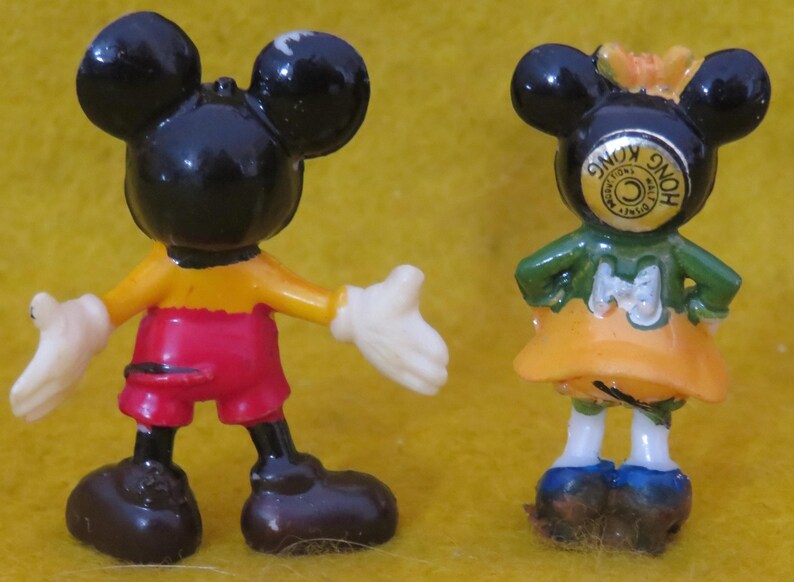 May include: Two small, vintage Mickey Mouse and Minnie Mouse figurines. Mickey is in red shorts, a yellow shirt, and white gloves. Minnie wears a green top and orange skirt. Both have black ears.