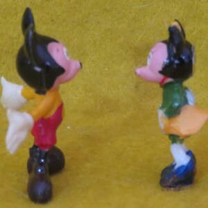 May include: Two small figurines of cartoon characters. One figure is wearing a yellow shirt, red shorts, black shoes, and white gloves. The other figure is wearing a green shirt, orange skirt, blue shoes, and a red bow tie. Both figures have black ears and pink faces.