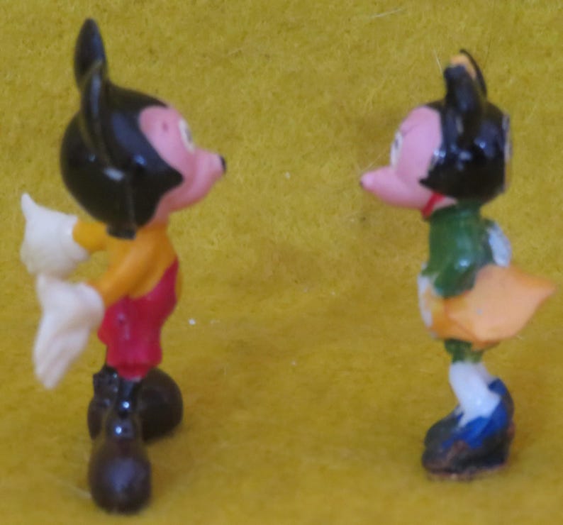 May include: Two small figurines of cartoon characters. One figure is wearing a yellow shirt, red shorts, black shoes, and white gloves. The other figure is wearing a green shirt, orange skirt, blue shoes, and a red bow tie. Both figures have black ears and pink faces.