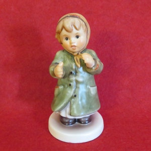 May include: A porcelain figurine of a young girl wearing a green coat and a brown hat. She has blonde hair and is standing on a white base.