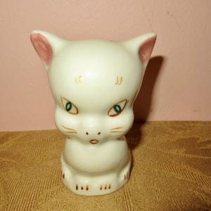 May include: A white ceramic cat figurine with pink ears and green eyes. The cat is sitting upright with its paws tucked underneath it.