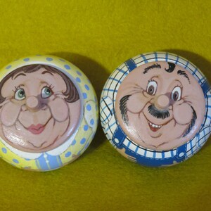 Puede incluir: Dos adornos redondos de madera con caras pintadas. El adorno de la mujer es amarillo con lunares azules y el adorno del hombre es azul con rayas blancas.