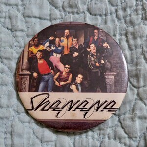 May include: A round button featuring a group of men in various outfits, with the word "Sha Na Na" in a stylized script. The image has a vintage aesthetic, with the band members posing in front of a brick wall.