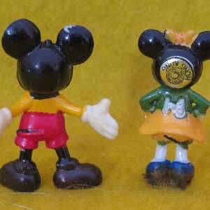 May include: Two small Mickey Mouse and Minnie Mouse figurines. Mickey is wearing red shorts, a yellow shirt, and white gloves. Minnie is wearing a green top, orange skirt, and blue shoes. Both have black ears and are standing on a yellow surface.