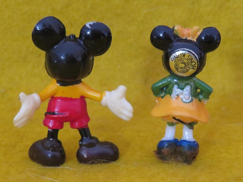 May include: Two small Mickey Mouse and Minnie Mouse figurines. Mickey is wearing red shorts, a yellow shirt, and white gloves. Minnie is wearing a green top, orange skirt, and blue shoes. Both have black ears and are standing on a yellow surface.