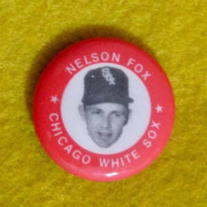 May include: A red button with a white circle in the center. The circle has a black and white photo of a baseball player wearing a Chicago White Sox hat. The text "NELSON FOX CHICAGO WHITE SOX" is printed around the circle.