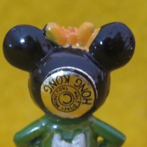 May include: A close-up of a vintage Mickey Mouse figurine. The figurine has black ears, a green body, and a yellow bow. A gold sticker on the front reads "Hong Kong" and "Walt Disney Productions".