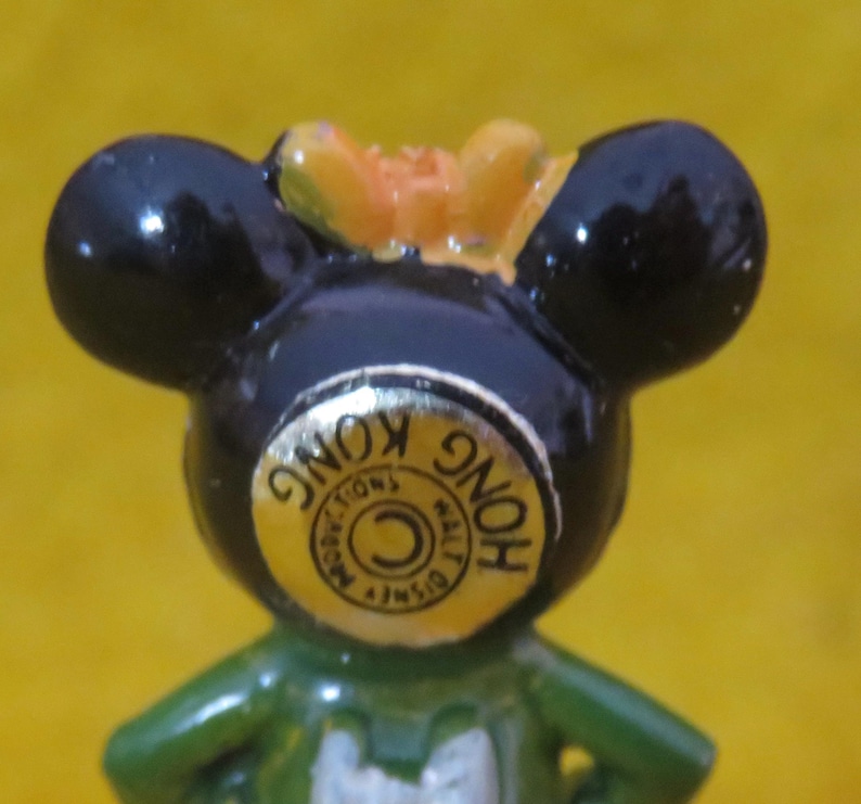 May include: A close-up of a vintage Mickey Mouse figurine. The figurine has black ears, a green body, and a yellow bow. A gold sticker on the front reads "Hong Kong" and "Walt Disney Productions".