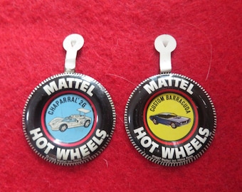 May include: Two round, black and silver Hot Wheels pinback buttons. The left button features a blue and white race car with the text "Chaparral '26" on a blue background. The right button features a yellow and blue car with the text "Custom Barracuda" on a yellow background. Both buttons have the text "Hot Wheels" in a red and white arc around the bottom.
