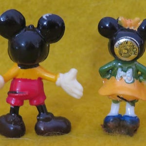 May include: Two small Mickey Mouse and Minnie Mouse figurines. Mickey is wearing red shorts, a yellow shirt, and white gloves. Minnie is wearing a green top, orange skirt, and blue shoes. The figurines are black, red, yellow, white, and blue.