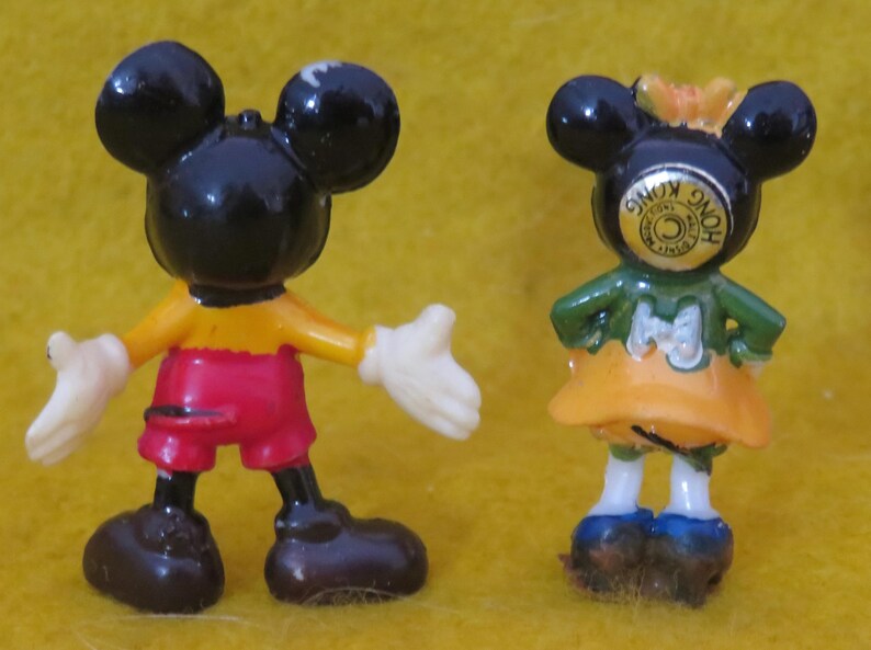 May include: Two small Mickey Mouse and Minnie Mouse figurines. Mickey is wearing red shorts, a yellow shirt, and white gloves. Minnie is wearing a green top, orange skirt, and blue shoes. The figurines are black, red, yellow, white, and blue.