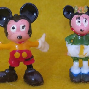 May include: Two small, vintage Mickey Mouse and Minnie Mouse figurines. Mickey is in a yellow and red outfit with black ears and shoes. Minnie is in a green top, white skirt, and blue shoes, with a bow on her head. Both have pink faces.